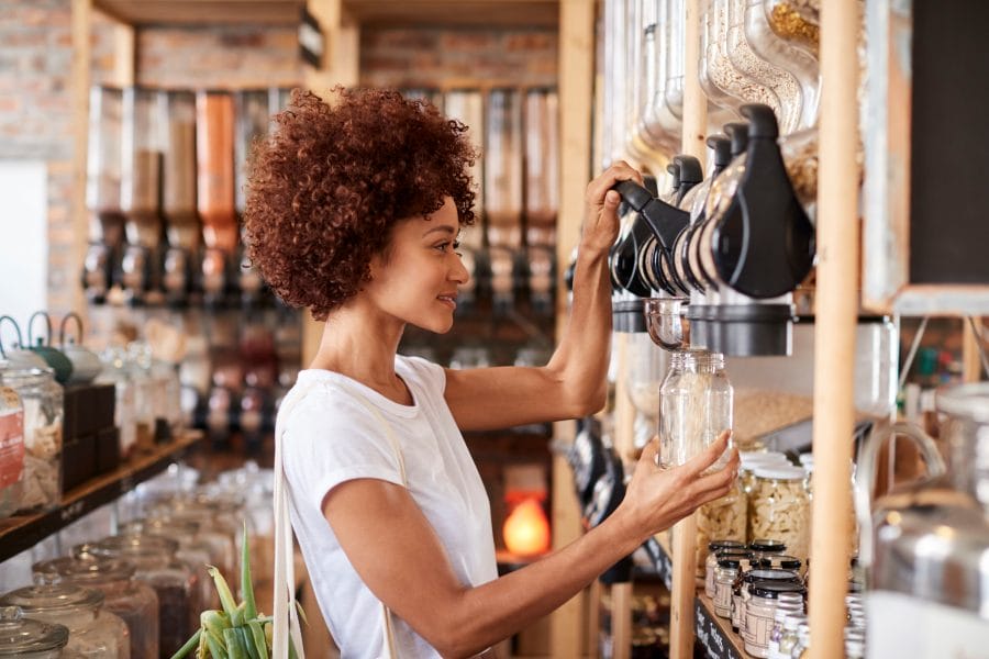 A woman is in a sustainable, plastic-free refill shop surrounded by various types of grain and cereals, showing how we can reduce waste. She is wearing a white t-shirt and holding up a glass jar, filling it with grains.