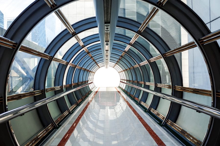 Photograph of a tunnel made from glass and metal leading to a bright light. Tunnel is surrounded by commercial buildings in the city of London