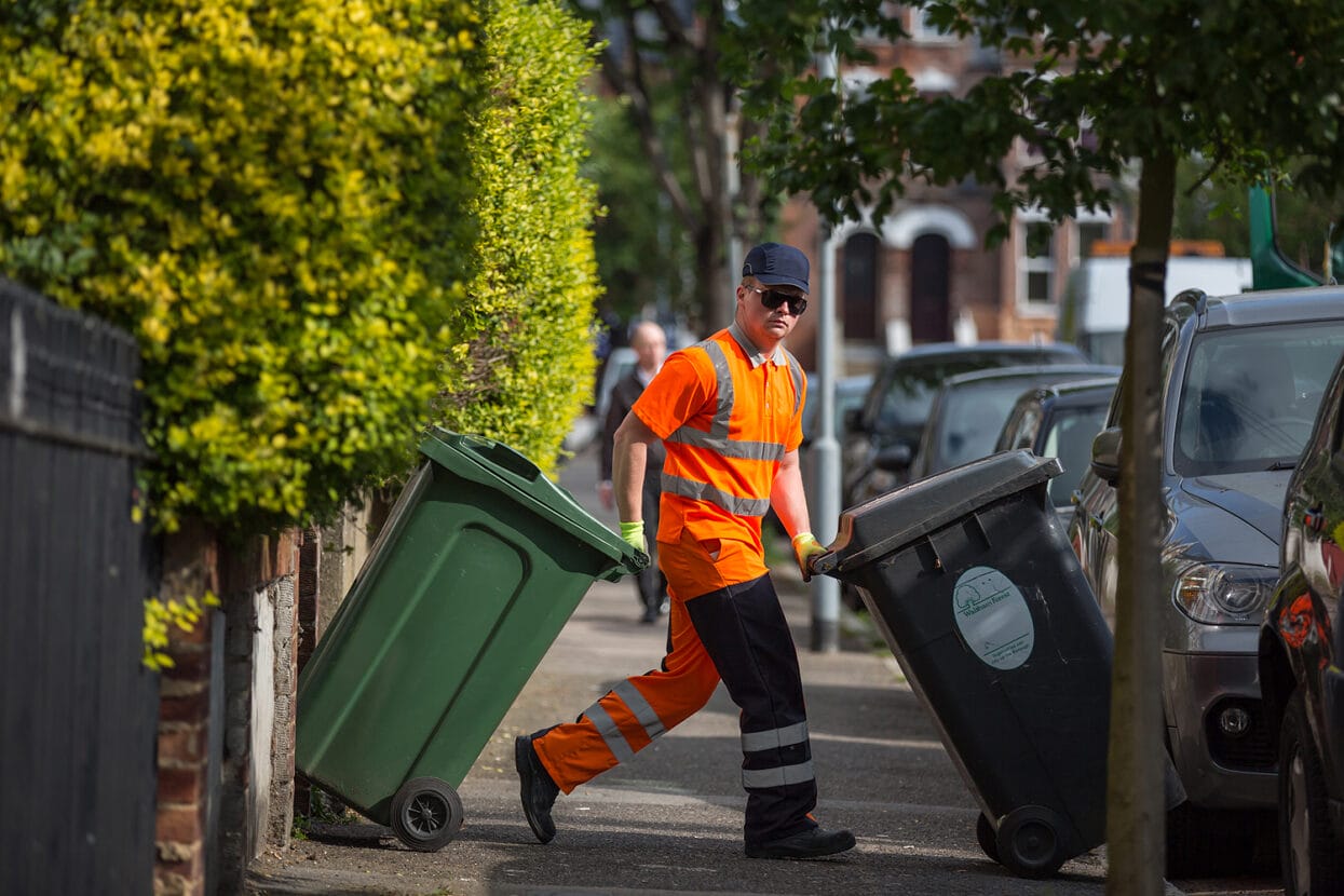 Waste collection employee in sunglasses, looking directly at the camera moving rubbish and recycling bins